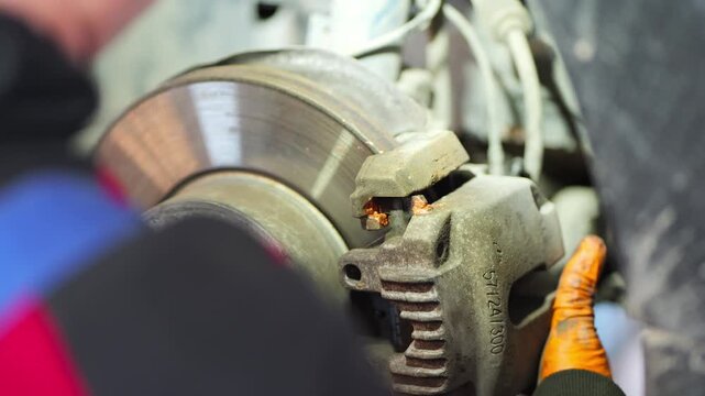 A close-up video shows a mechanic wearing orange gloves servicing a car&rsquo;s front brake system, carefully positioning the brake caliper over the disc and applying lubricant to ensure smooth 