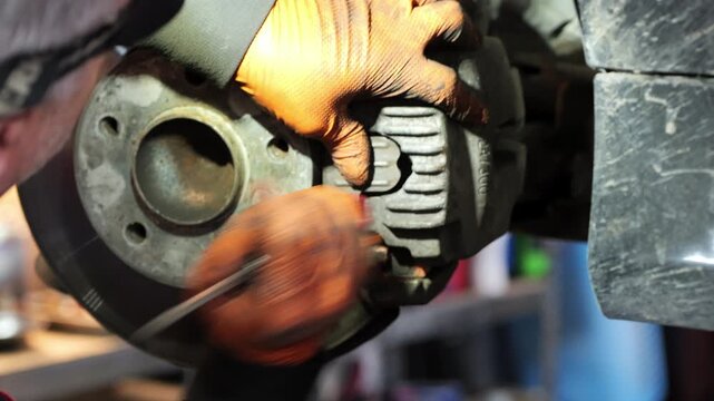 A close-up video shows a mechanic wearing orange gloves servicing a car&rsquo;s front brake system, carefully positioning the brake caliper over the disc and applying lubricant to ensure smooth 