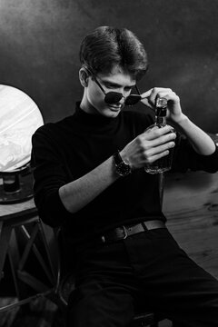 A stylish black and white portrait of a young man wearing dark sunglasses and a black turtleneck. He is holding and examining a glass bottle, with a globe visible in the background. 