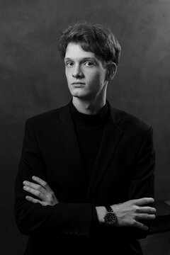 Classic black and white portrait of a thoughtful young man with freckles, wearing a black blazer and turtleneck. The subject is posing with crossed arms against a dark textured background.