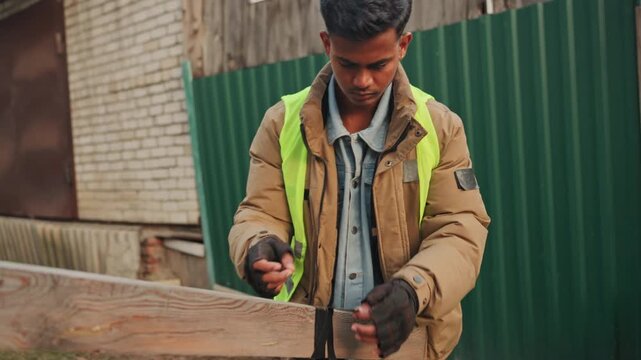 Man repairing wooden fence outdoors wearing highvisibility jacket and gloves, measuring and fixing plank with hammer and screwdriver against brick wall focused daytime urban repair project emphasizing