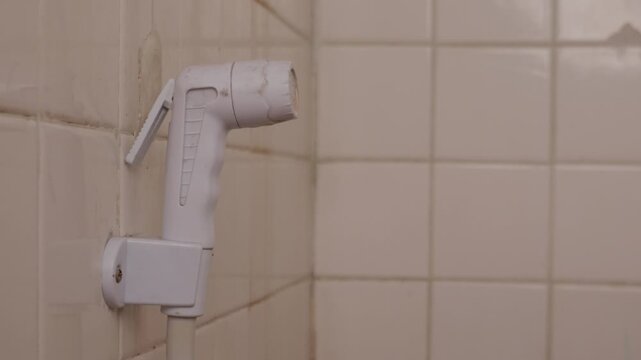 White plastic bidet hand shower sprayer mounted on clean beige square ceramic tile bathroom wall with white hose