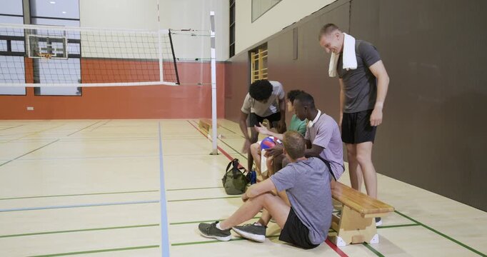 Diverse men in jerseys greeting arriving teammate, passing volleyball on bench on break, copy space