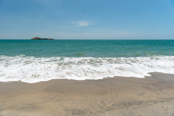 Obraz premium Turquoise waves lap a Pacific ocean sandy shore toward the horizon where a small tropical island rests beneath a clear blue sky, serene and untouched for a peaceful getaway - stock photo