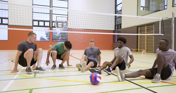 Diverse male volleyball team warming up in gym, third player falling, others stretching toward ball
