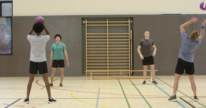 Male volleyball teammates sending and receiving volleyballs from high toss in school gym, training