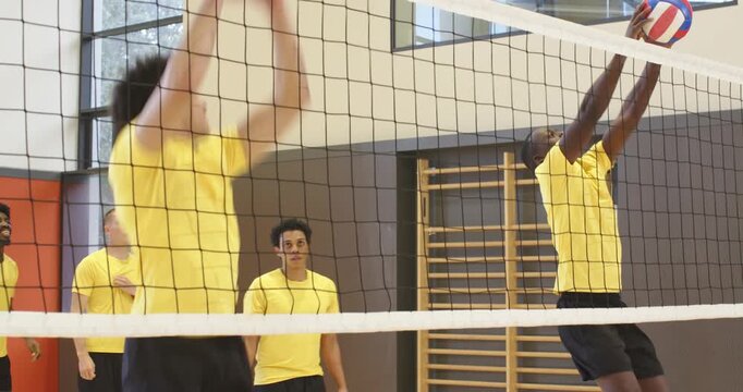 Diverse male team in yellow passing blocking at net after toss with volleyball drilling school gym