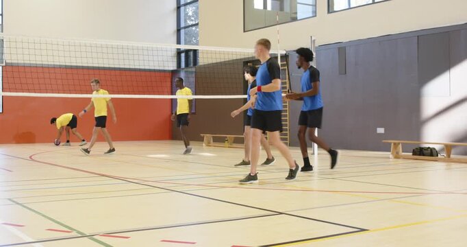 Diverse male blue team reacting to yellow serve, receiving at net, jumping to score at gym
