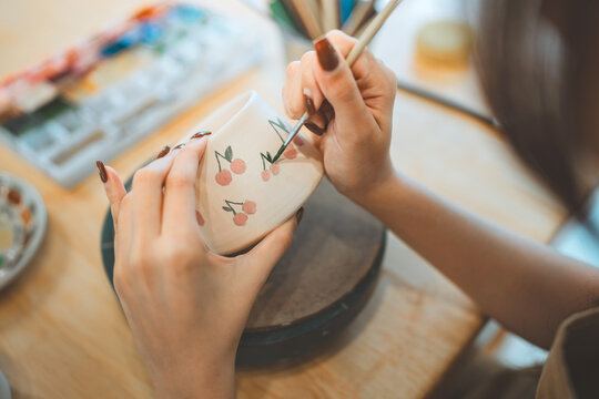 Close up of an artist's hands painting cherry patterns on a ceramic bowl in a studio. Concept of mindfulness, handmade craft, and creative hobby.