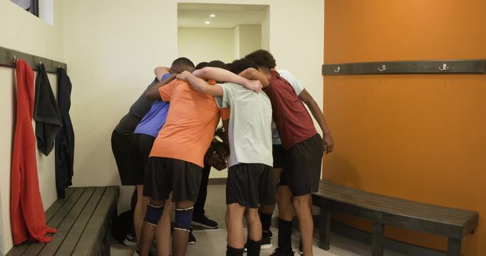 Diverse male team hearing kneeling coach huddling chanting preparing in locker rm with volleyball