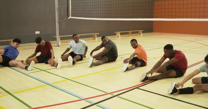 Diverse male team following coach from left, seated stretching hamstrings and warming up beside net