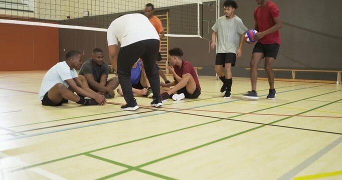 Diverse male team listening, coach gesturing at gym, players jogging with volleyballs learning