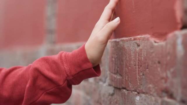 Black woman inspects old red brick, hand glides along crumbling mortar, red sweater sleeve visible, closeup on surface erosion and patina, patient deliberate movement and quiet restorative curiosity.