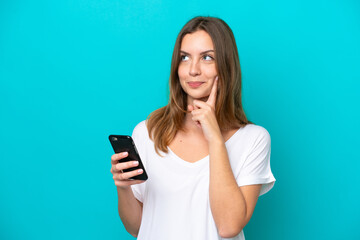 Fototapeta premium Young caucasian woman isolated on blue background using mobile phone and thinking
