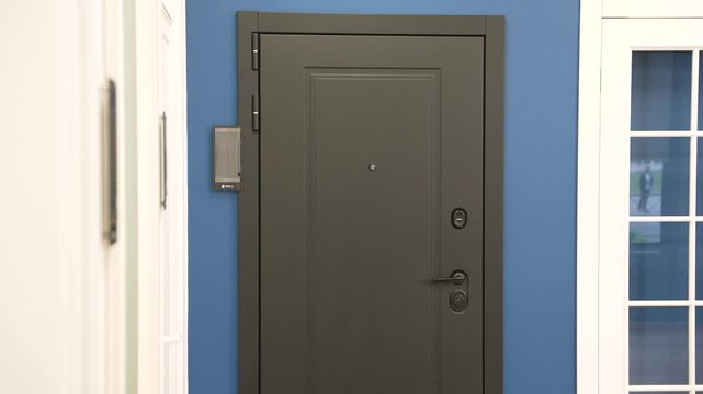 Smooth pan shot of a modern gray security door with a handle, peephole, and lock. The door is set in a blue wall in a showroom or apartment hallway. A high tech security system is nearby