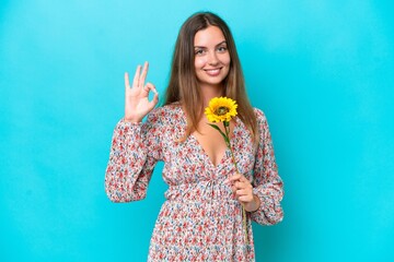 Young caucasian woman holding sunflower isolated on blue background showing ok sign with fingers