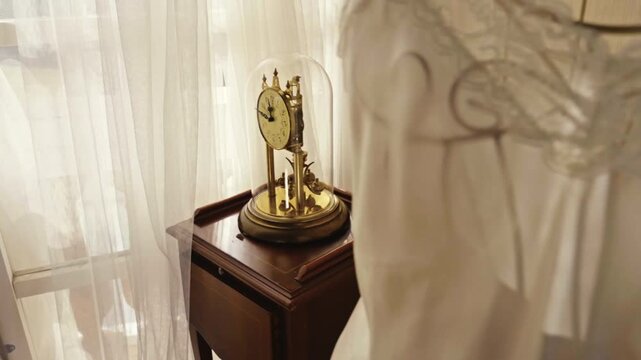 Vintage brass torsion clock under glass dome on wooden pedestal in bright interior with white curtains