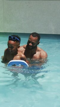 Vertical video: Guiding dad holding kickboard, teaching son in trunks kicking in pool, green ring