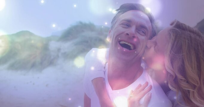 Climbing woman kissing cheek, man laughing, sharing love on sandy dunes, white-tee, pink-bokeh