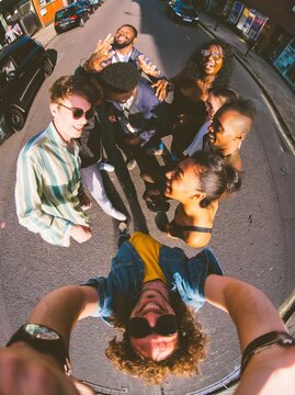 Group of young adults posing together in a circle on a city street, captured with a wide angle fisheye lens perspective, showcasing vibrant urban surroundings