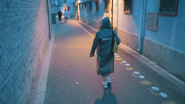 Hooded woman walking down narrow alley carrying yellow bag and wearing long coat, illuminated by warm street lamps and cool twilight, painted pavement markers and brick walls, slow solitary pace