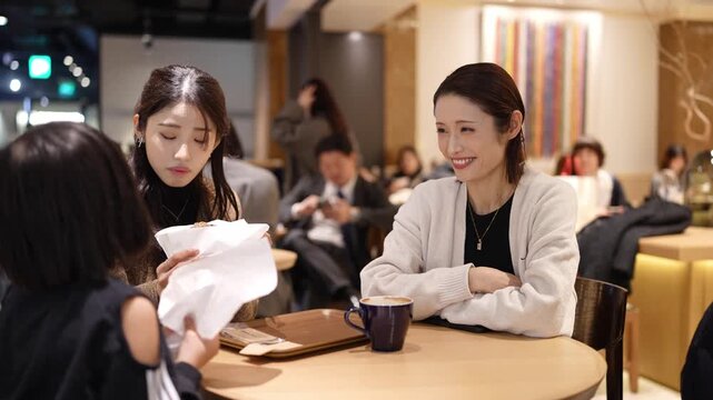 Three women converse over coffee in a cozy cafe. One smiles warmly, engaged in conversation. Another focuses on documents, appearing thoughtful. The setting feels relaxed, filled with soft lighting
