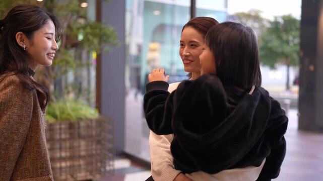 Three women converse in a modern urban setting. One gestures while speaking, showing engagement. Another smiles warmly, indicating a friendly mood. The third listens, with a soft, pleasant expression