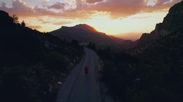 Drone view of a runner in a red shirt moving along a quiet road. Golden sunset rays, mountains and motion create a sense of freedom, endurance and peaceful solitude.