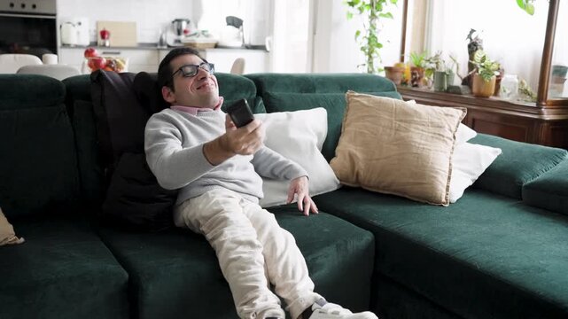 Man with a congenital condition relaxing on a comfortable couch in his living room. He holds a remote control, smiling and enjoying a movie