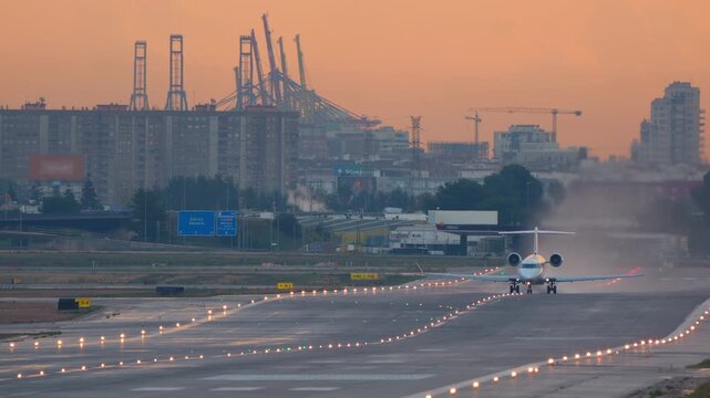 A passenger airplane accelerates down a lit runway and takes off against a hazy city skyline during a beautiful orange sunset. The port cranes are visible in the background