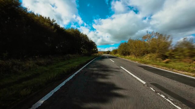 Driving along countryside highway with left hand traffic in United Kingdom under cloudy blue sky POV. Car moving through rural British road surrounded by fields and hedges first person view. SUV