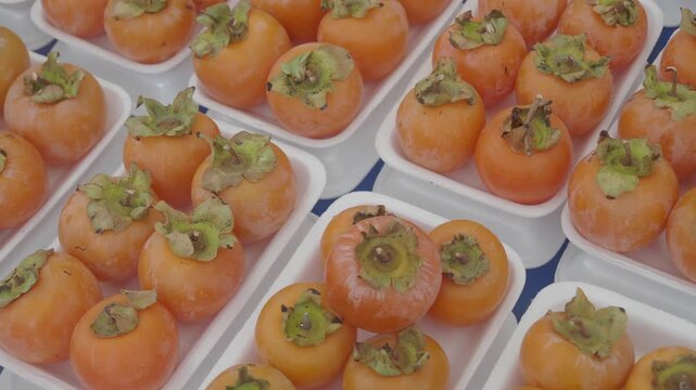 Rows of fresh persimmons in plastic trays displayed at market stall motion.
