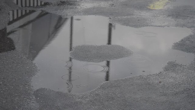 Raindrops falling into asphalt puddle reflecting overcast sky and railing.