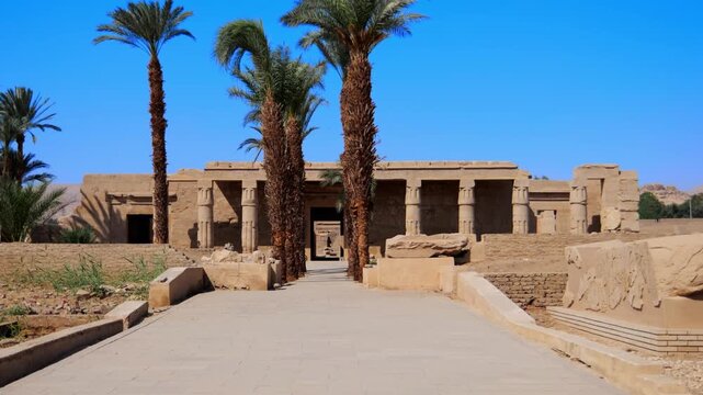 Seti I mortuary temple entrance with palm tree alley and ancient stone ruins in Luxor Egypt. Scenic pathway under vivid blue sky.