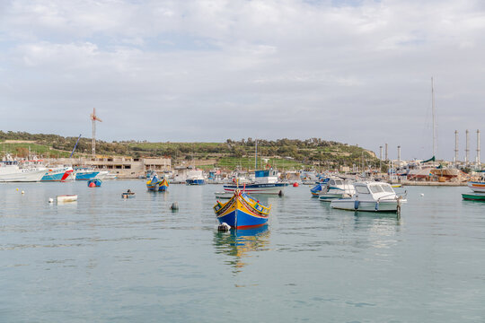 View of vibrant, colorful boats bobbing gently on the tranquil, turquoise waters of the harbor against a backdrop of buildings, Marsaxlokk, Malta.