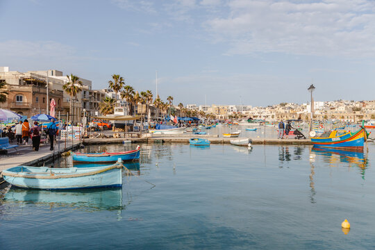 View of vibrant fishing boats bobbing on the tranquil, cerulean harbor waters, framed by bustling quayside and sun-kissed buildings, Marsaxlokk, Malta.