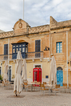 View of old buildings with blue doors, a red telephone box, and tables with umbrellas standing on the square, Marsaxlokk, Malta.