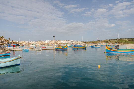 View of vibrant, colorful fishing boats bob gently on the tranquil, deep blue water under a soft, cloudy sky, Marsaxlokk, Malta.