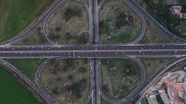 Aerial view of a busy highway interchange with multiple lanes and vehicles moving along the roads, contrasting with the surrounding green fields, Rome, Lazio, Italy.