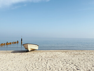 white wooden rowing boat rests on a bright sandy Baltic beach in clear morning light, with weathered wooden groynes reflecting in the glassy shallow water