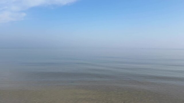 perfectly calm, glassy sea beneath a vast pale blue sky with wispy clouds, where the horizon dissolves into soft coastal mist, creating an exceptionally minimal and serene seascape