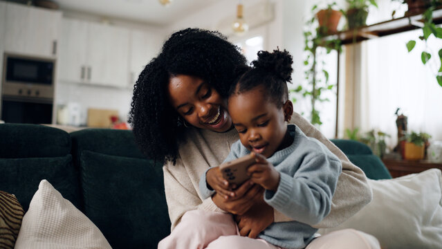African American mother and daughter sitting on sofa, laughing and bonding while using a smartphone together at home