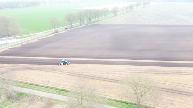 High angle drone footage showing a red tractor steadily plowing a large agricultural field from left to right. As the plow turns the soil, a flock of white birds swarms the freshly overturned earth