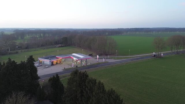 A cinematic drone shot capturing a roadside service station and automated car wash facility during the late afternoon. The warm low sun casts long shadows across the asphalt, a white van drives by