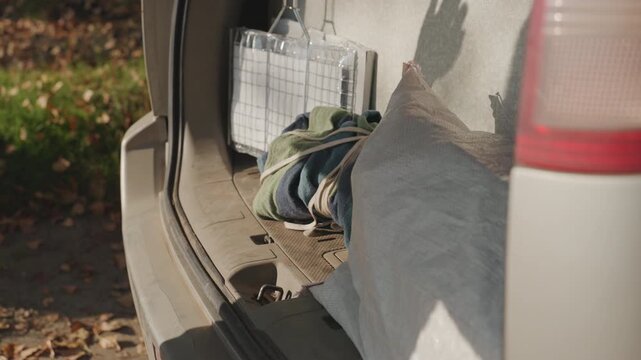 Car trunk with blankets pillows and gear, hand arranging equipment in open hatch, green sweater sleeve visible, soft autumn light and fallen leaves, closeup of cargo area showing straps, cushions