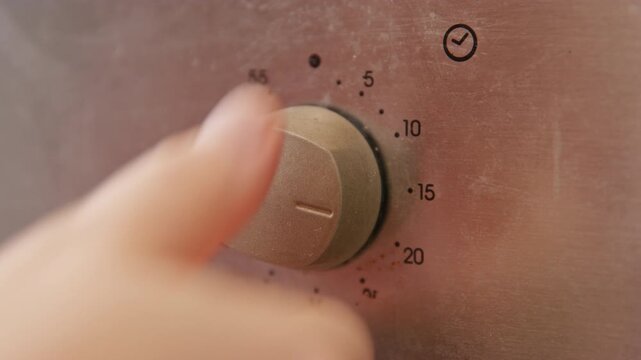 Close-up of a woman hand in a red sleeve turning a metallic oven timer knob with minute markings, kitchen appliance detail, home cooking routine, meal preparation moment, domestic lifestyle scene.