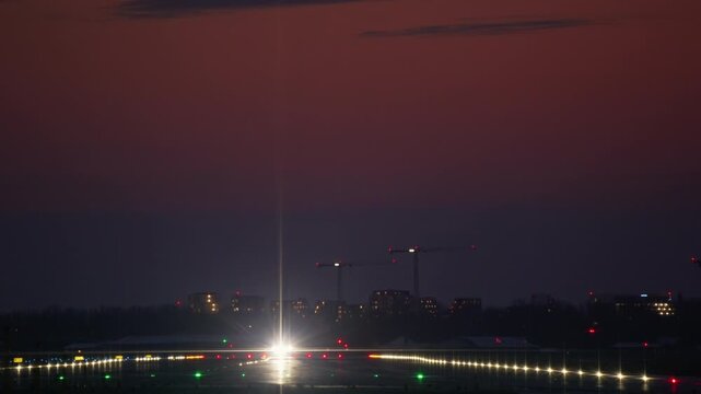 A passenger airplane with bright lights accelerates down an illuminated runway and takes off into the twilight sky. A city skyline with cranes is visible in the background