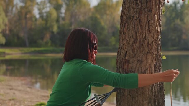Woman sets hammock. Person prepares relaxing outdoor rest. Women stake out peaceful resting spot with careful knotting. Woman expertly secures hammock beside serene lakeside for leisure