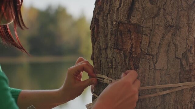 Closeup white woman tying rope tree by lakeside, hands skillfully knotting natural fiber cord around rough bark, preparing secure anchor for hammock, autumn light casting warm tones over calm water,