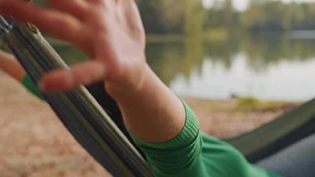 Woman lounging in hammock by lake, camping relaxation with gentle stretch green sweater and sunglasses, calm water reflections on shoreline, soft autumn light, slow breath and reflective mood,